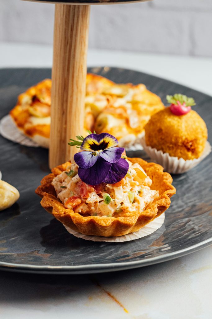 A close-up of gourmet tartlets garnished with a purple edible flower on a dark plate.